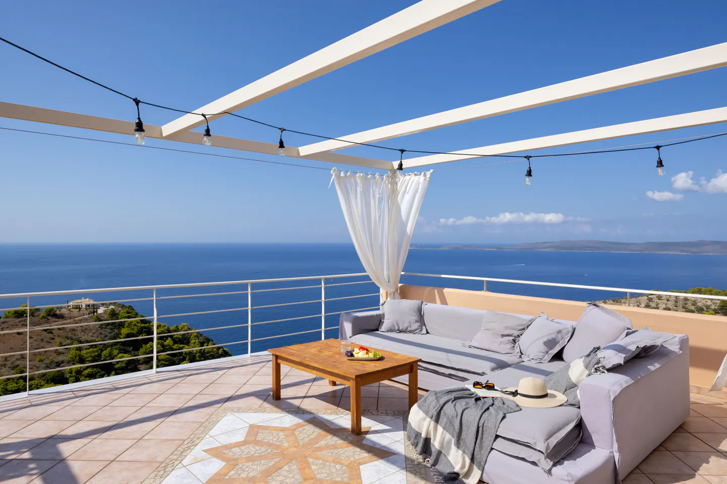 Outdoor patio with gray sofa, wooden table, and ocean view. String lights hang above.
