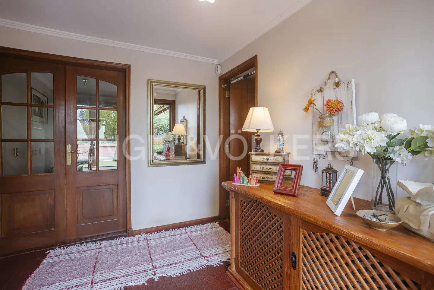 A home's entryway with a wooden console table, lamp, flowers, and a mirror. A red and white rug lies on the floor.