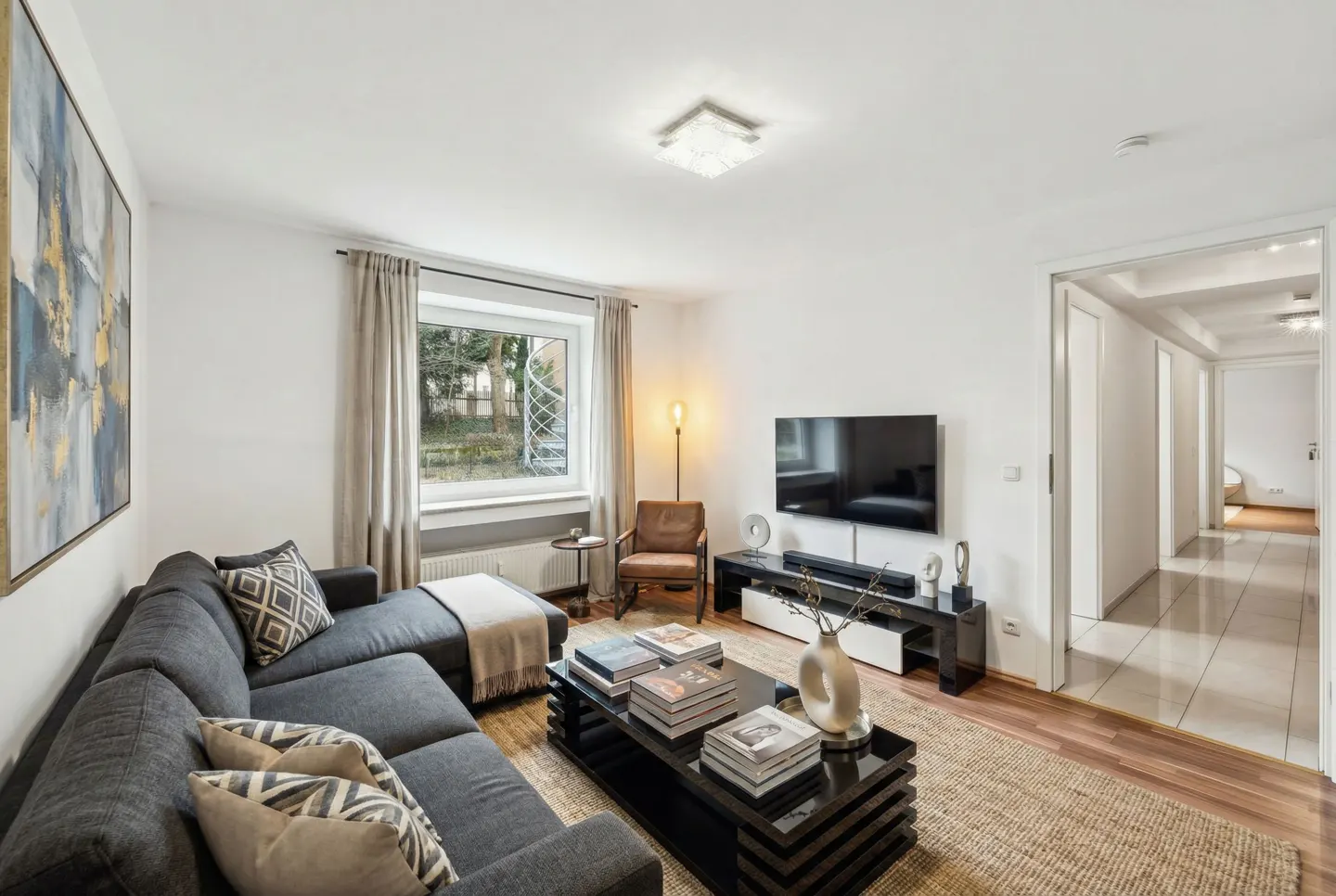 Living room with gray sectional sofa, black coffee table with books, TV, and hallway view. Neutral tones and modern decor.