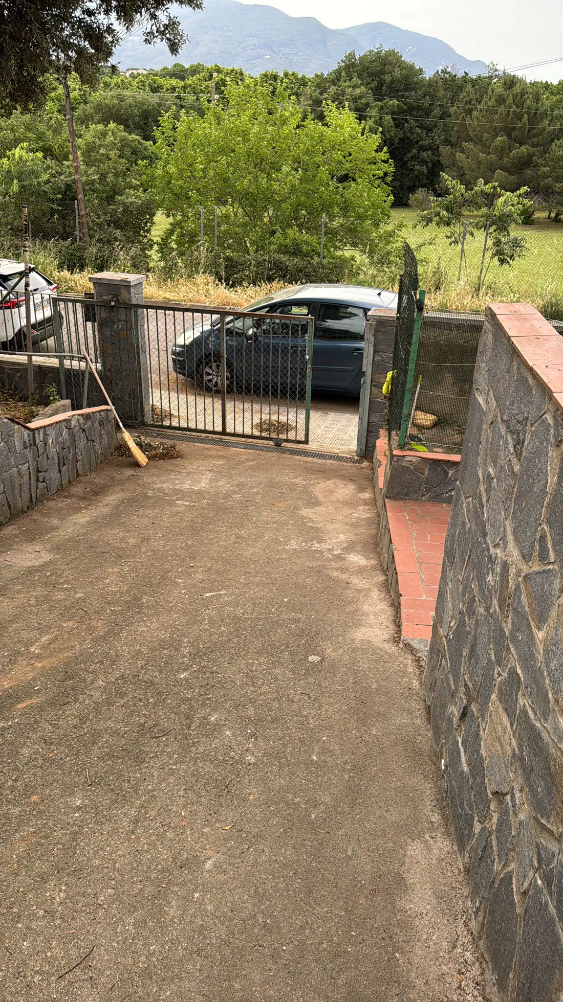 A concrete driveway leads to a black metal gate, with a blue car parked behind it. A broom leans against a stone wall to the left. Green trees are in the background.
