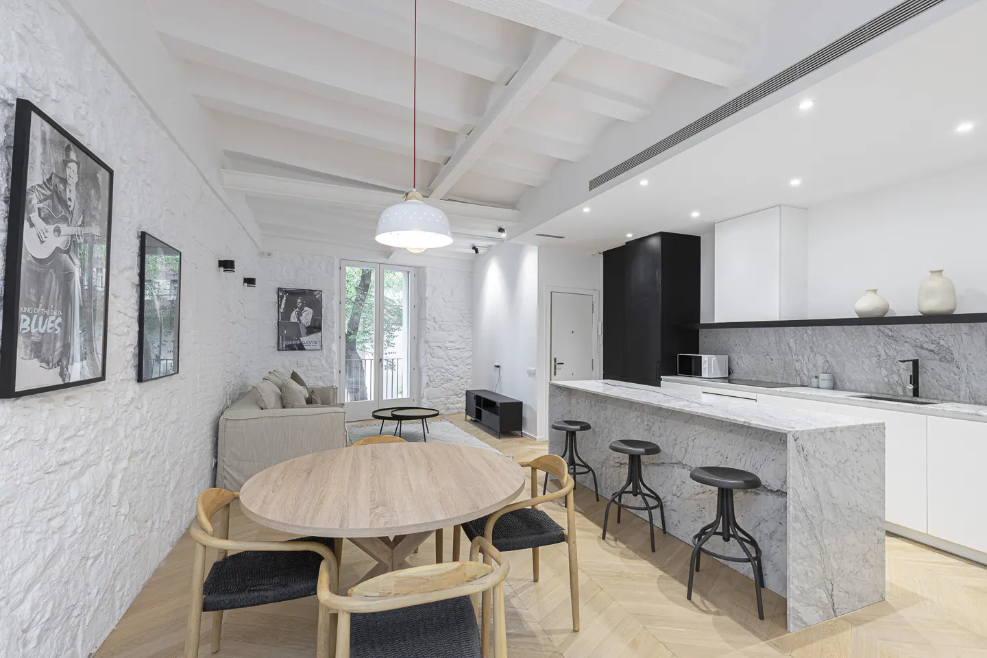 Bright, open-concept living space with white walls, wood floors, and a marble kitchen island with black stools. A round wood table sits in the foreground.