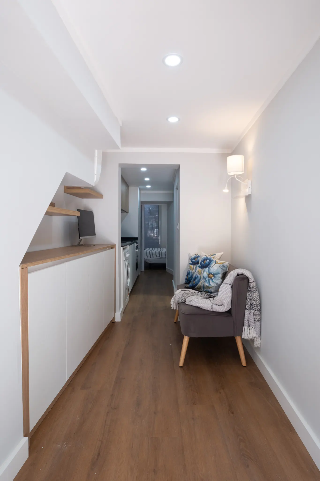Bright, modern apartment interior with wood floors, white walls, and recessed lighting. A gray chaise lounge sits near a wall lamp.