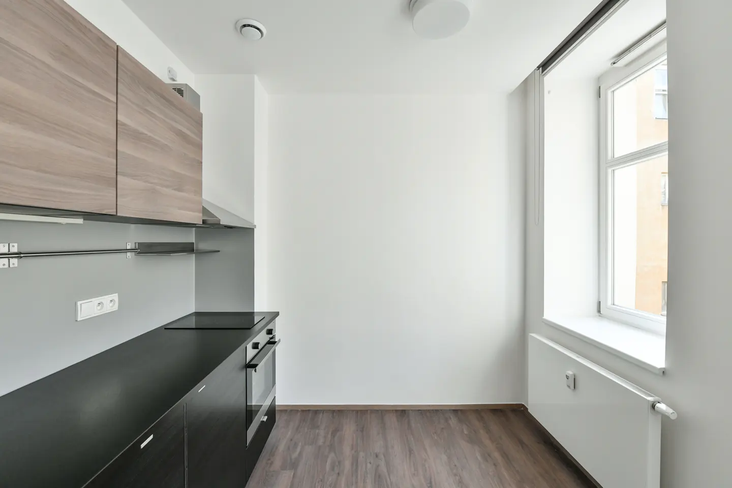Modern kitchen with wood-grain upper cabinets, black lower cabinets, a built-in oven, and a window with a white radiator.