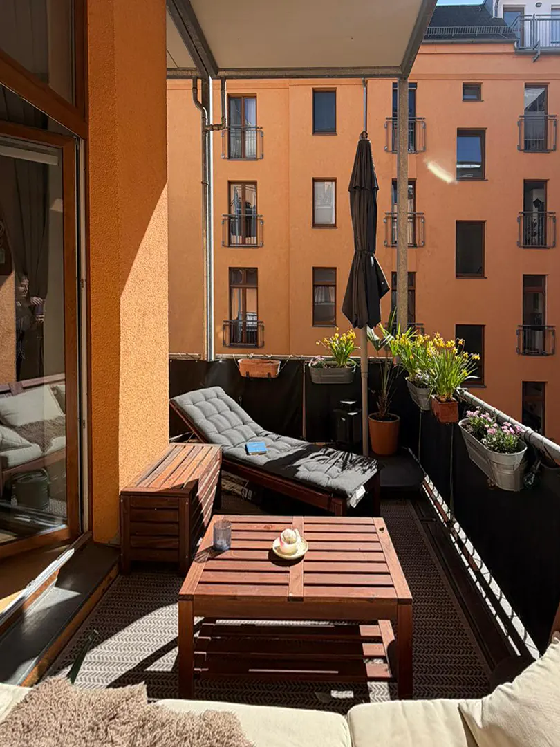 Sunny balcony with wooden furniture, including a table, bench, and lounge chair. Flowers in pots line the railing, with an orange building in the background.