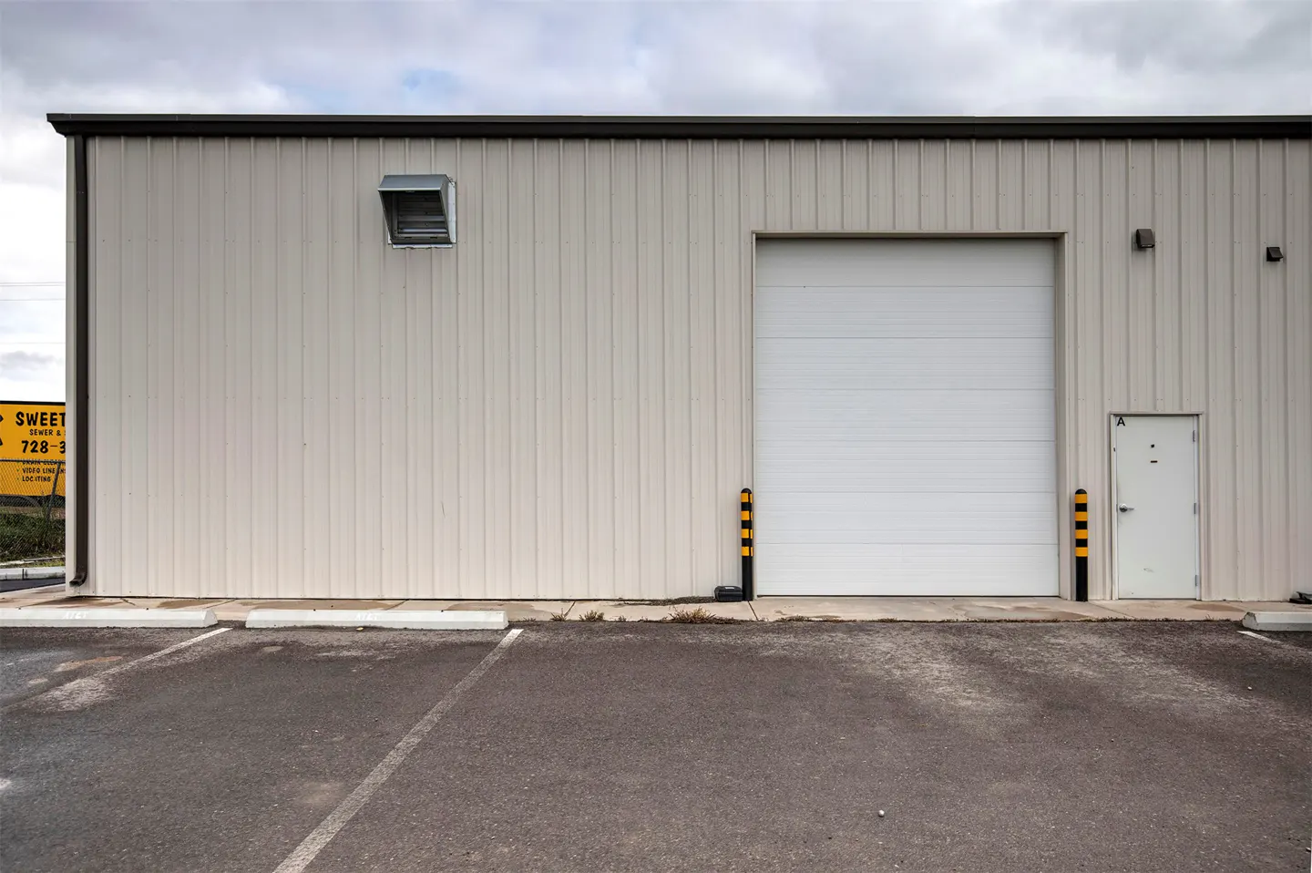 Exterior view of a beige metal warehouse with a large white roll-up door, a side entry door, and an asphalt parking lot.