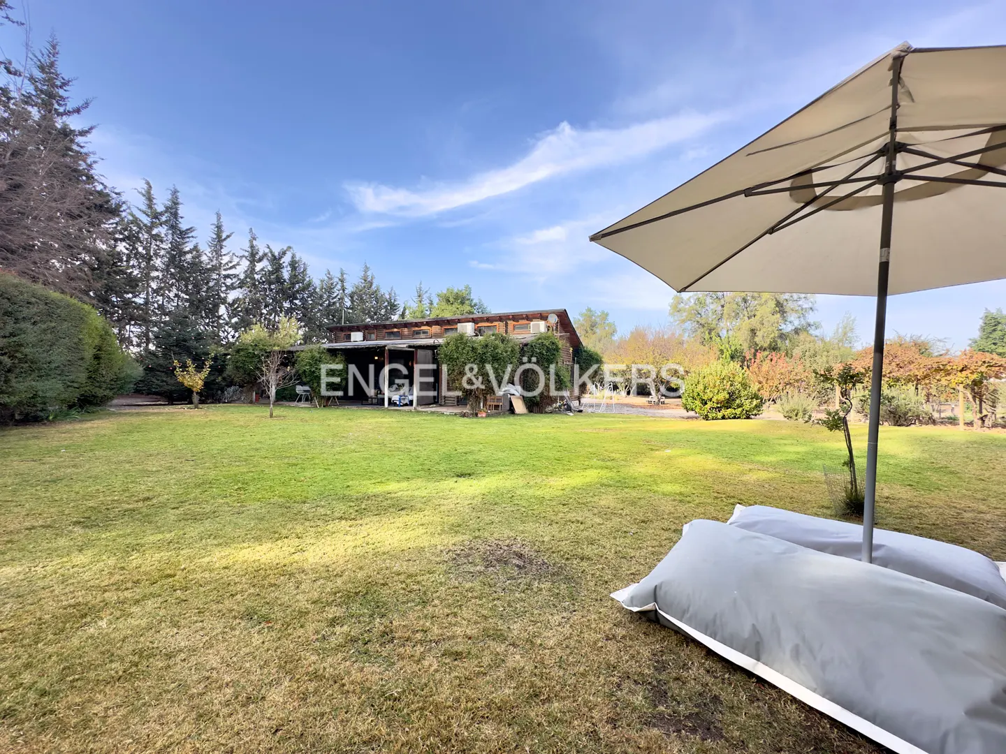 Large grassy backyard with a patio umbrella and bean bag chairs, facing a rustic house under a clear blue sky.