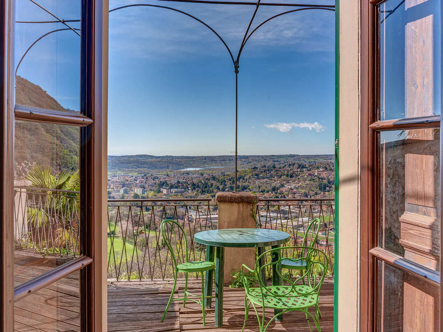 View from open doors to a balcony with green table and chairs overlooking a city and lake under a blue sky.