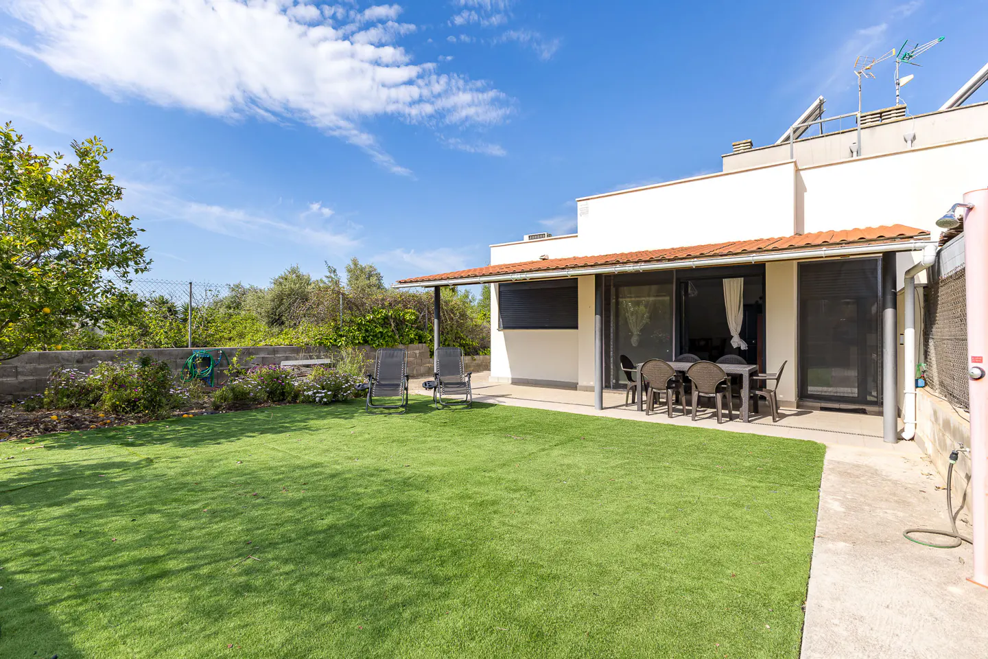 Backyard view of a white house with a red tile roof, green lawn, patio furniture, and blue sky.