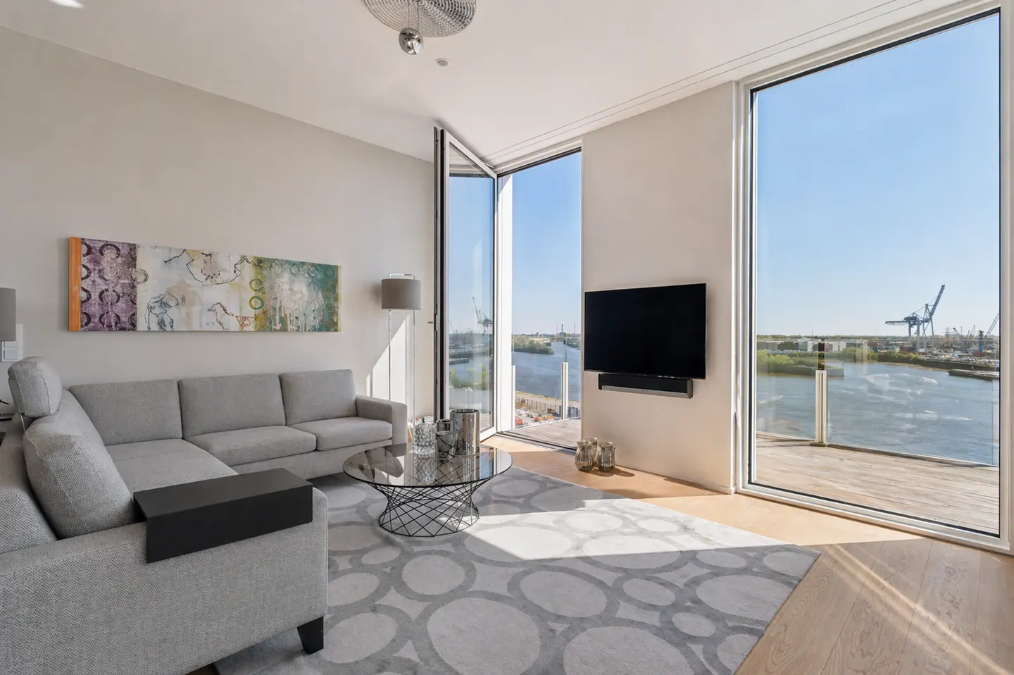Bright living room with gray sectional sofa, round glass table, and large windows overlooking a harbor with cranes. Abstract art hangs on the wall.