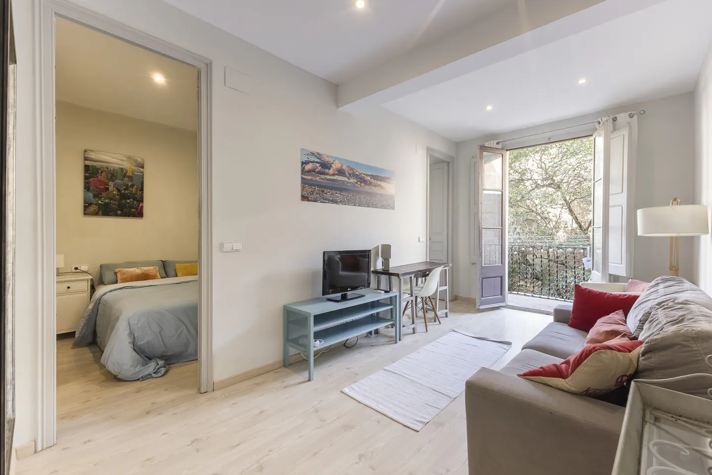 Bright living room with a grey sofa, blue TV stand, and a doorway leading to a bedroom with a bed and colorful art.