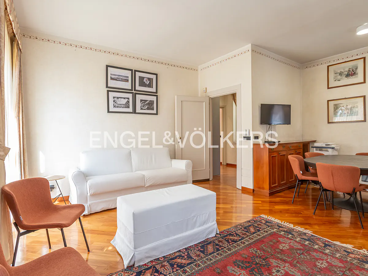 Living room with white sofa, ottoman, and brown chairs on a wood floor with a red and blue rug. A TV and dining table are in the background.