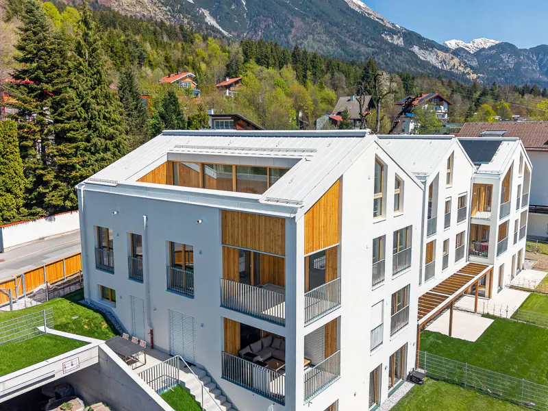 Aerial view of a modern white apartment building with wood accents, balconies, and a mountain backdrop.