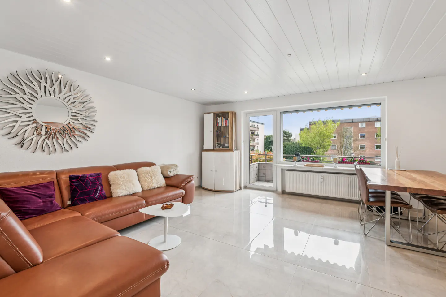 Bright living room with a brown leather sectional sofa, white walls, and a large window with a view of trees and buildings.