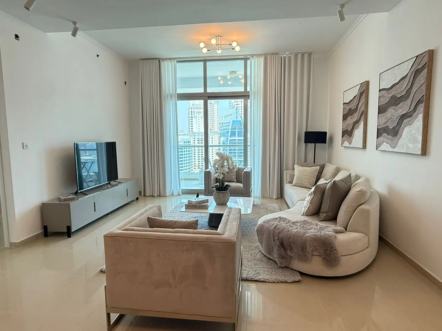Bright living room with beige sofas, a TV, and city views through large windows. Neutral tones and modern decor create a serene atmosphere.