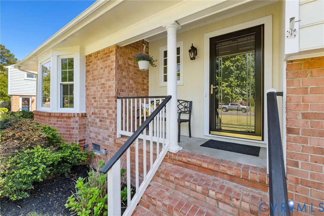 Front entrance of a brick home with a black door, white railing, and a small porch with a black chair.