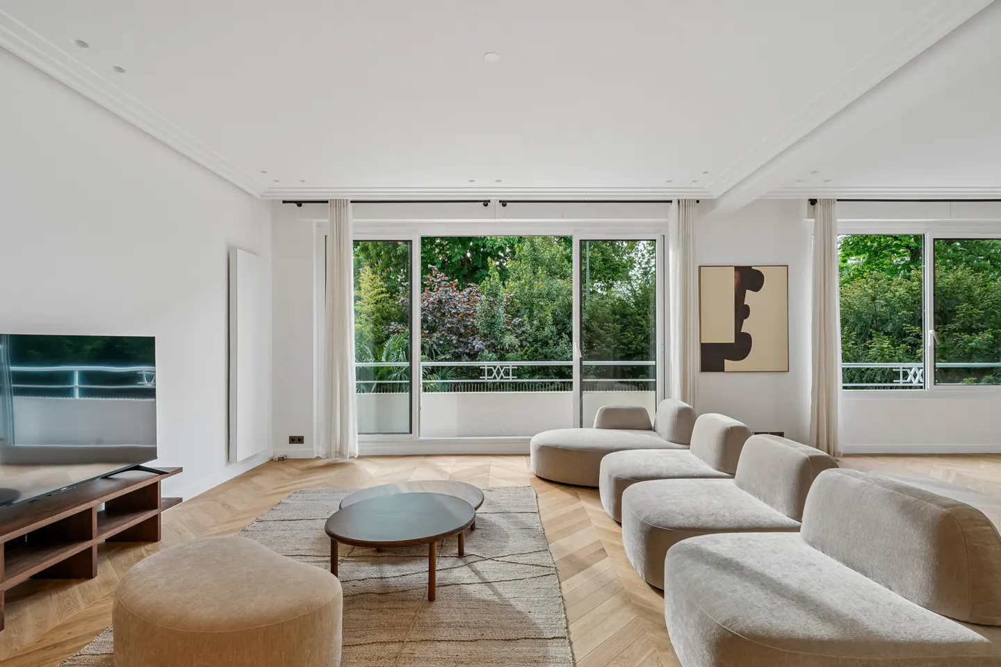 Bright living room with a beige sectional sofa, wood floors, and large windows overlooking lush greenery. A modern coffee table sits on a patterned rug.