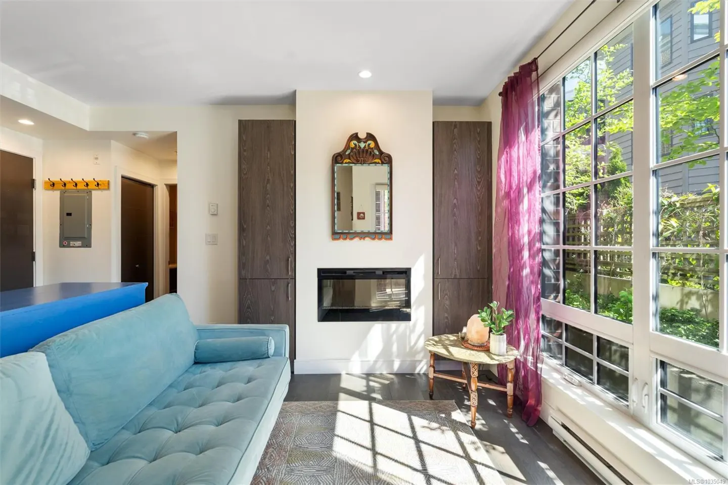 Living room with a blue sofa, fireplace, and large windows with purple curtains. A small table sits near the window.