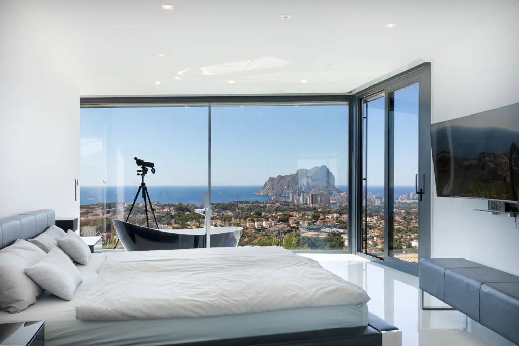 Bedroom with a large window view of the ocean, city, and mountain. A telescope and black tub are near the window. A bed and TV are in the room.