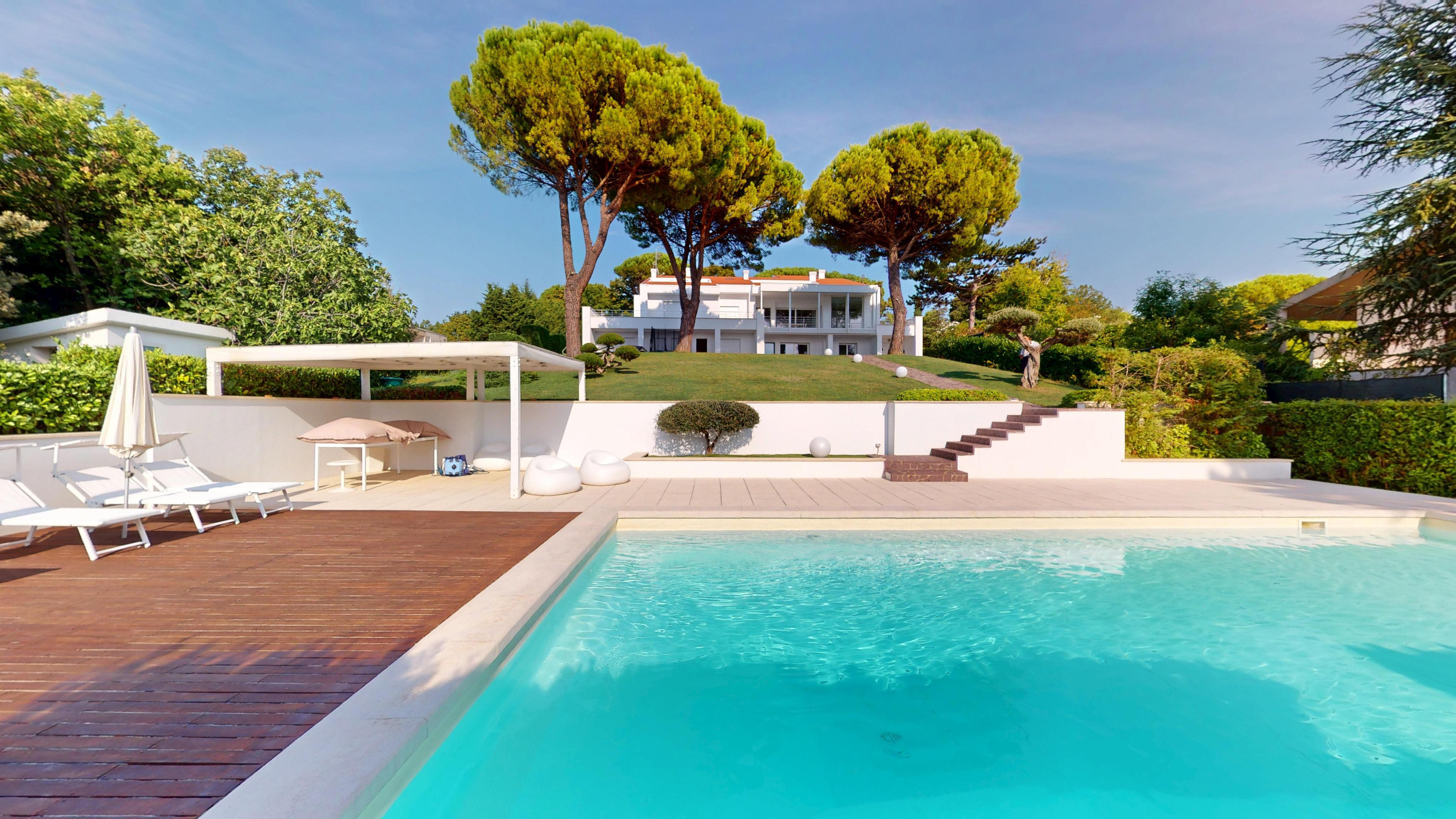 A bright, sunny view of a modern white house with a pool. Lounge chairs sit on a wooden deck next to the turquoise water.
