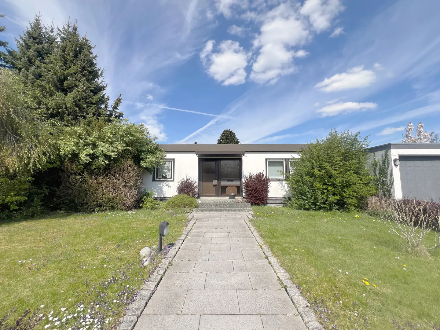 Exterior view of a one-story white house with a gray roof and a stone walkway leading to the front door. Green lawn and trees surround the house.