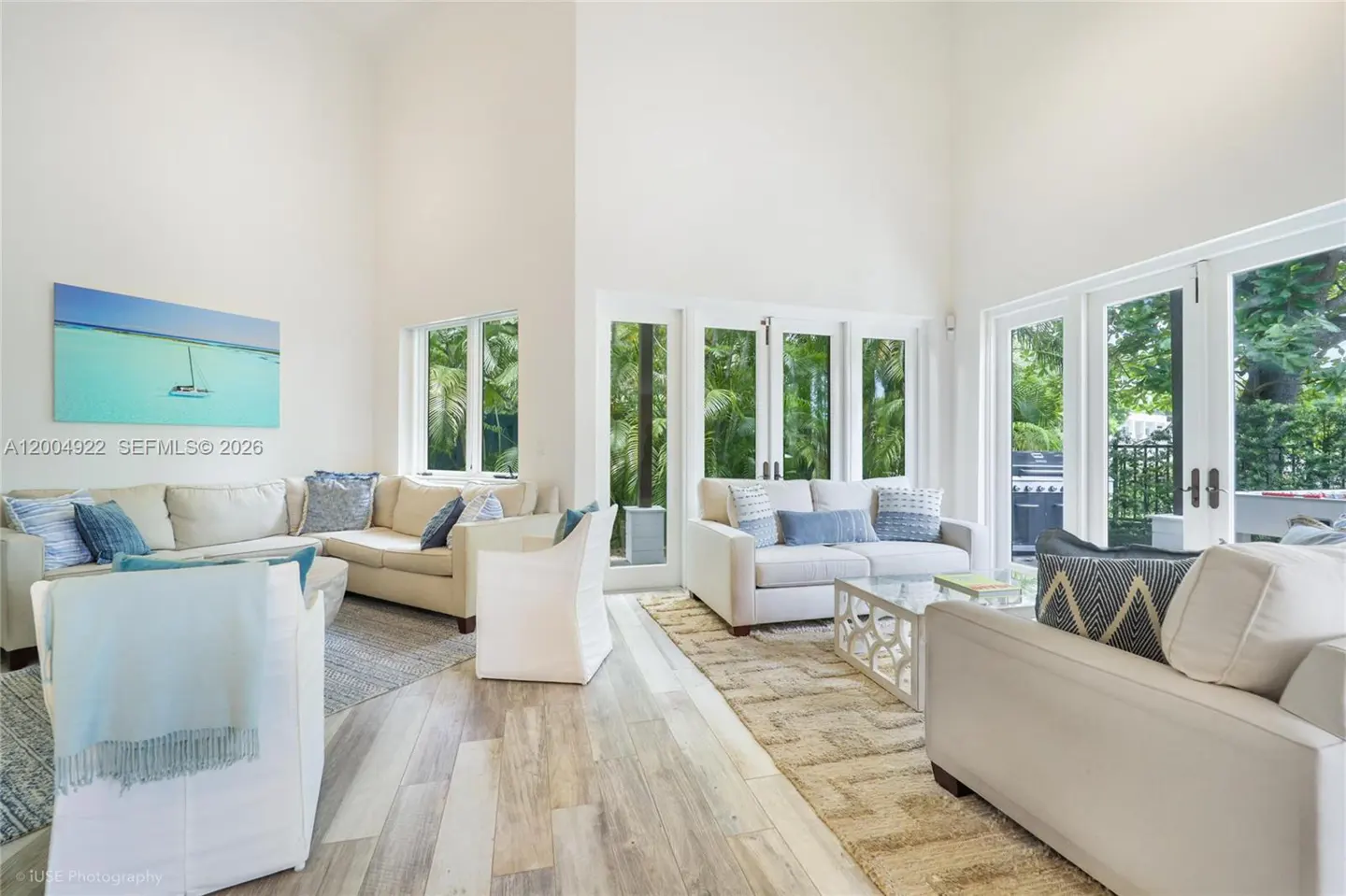 Bright living room with white sofas, blue pillows, and a painting of a sailboat. French doors lead to a lush green outdoor space.