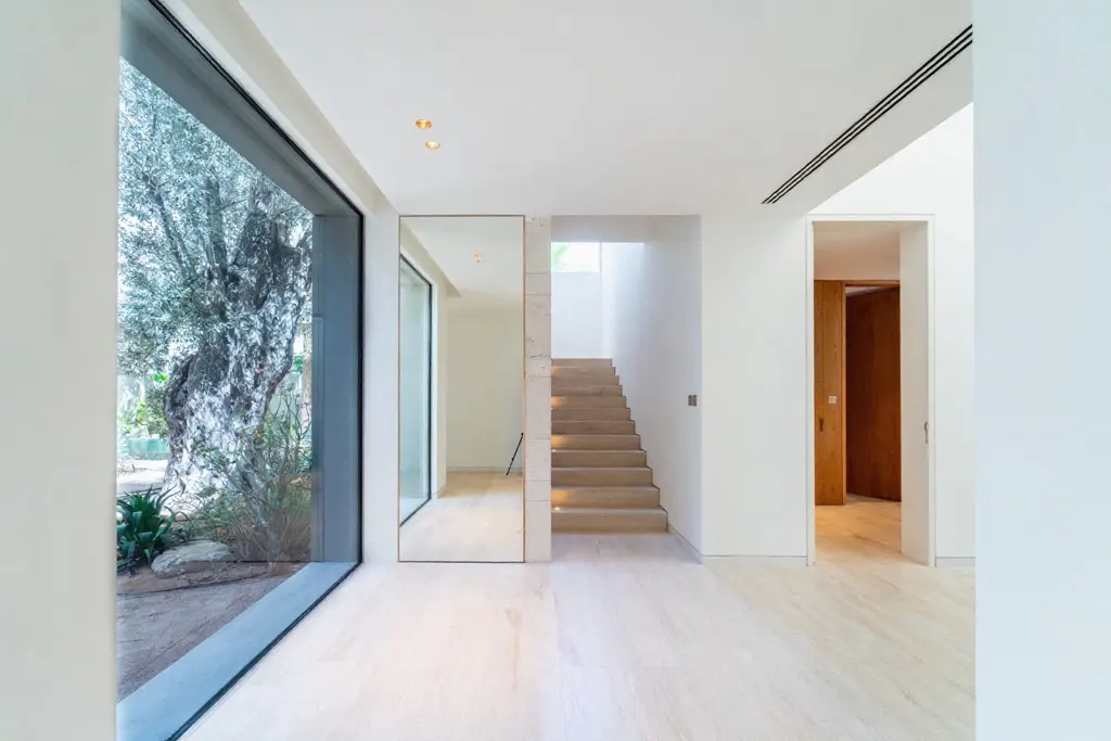 Bright, modern foyer with light wood floors, white walls, and a staircase. A large window shows a tree outside.