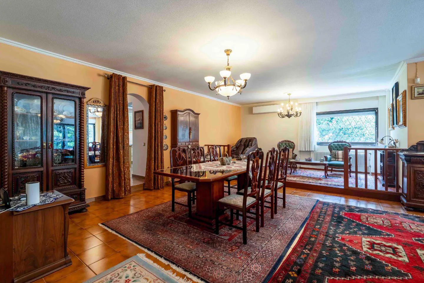Formal dining room with a wooden table, six chairs, ornate chandeliers, and traditional rugs on a tiled floor.