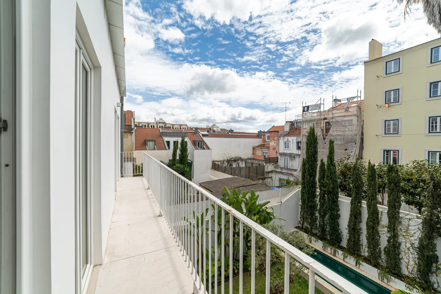 Balcony view with white railings overlooking a garden, buildings with red roofs, and a cloudy blue sky.