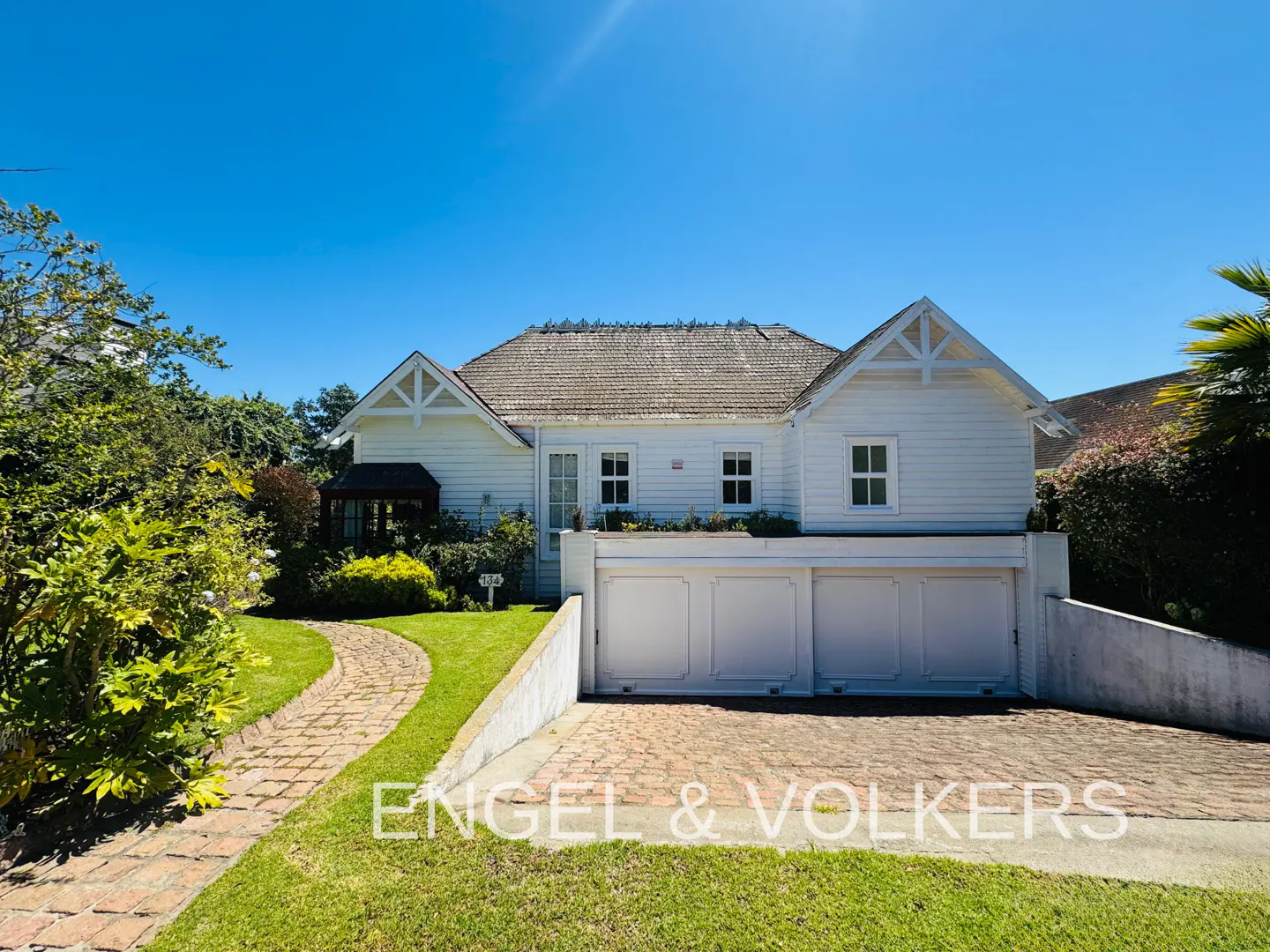 Exterior of a white house with a brick driveway and a blue sky. The house has a brown roof and white garage doors.