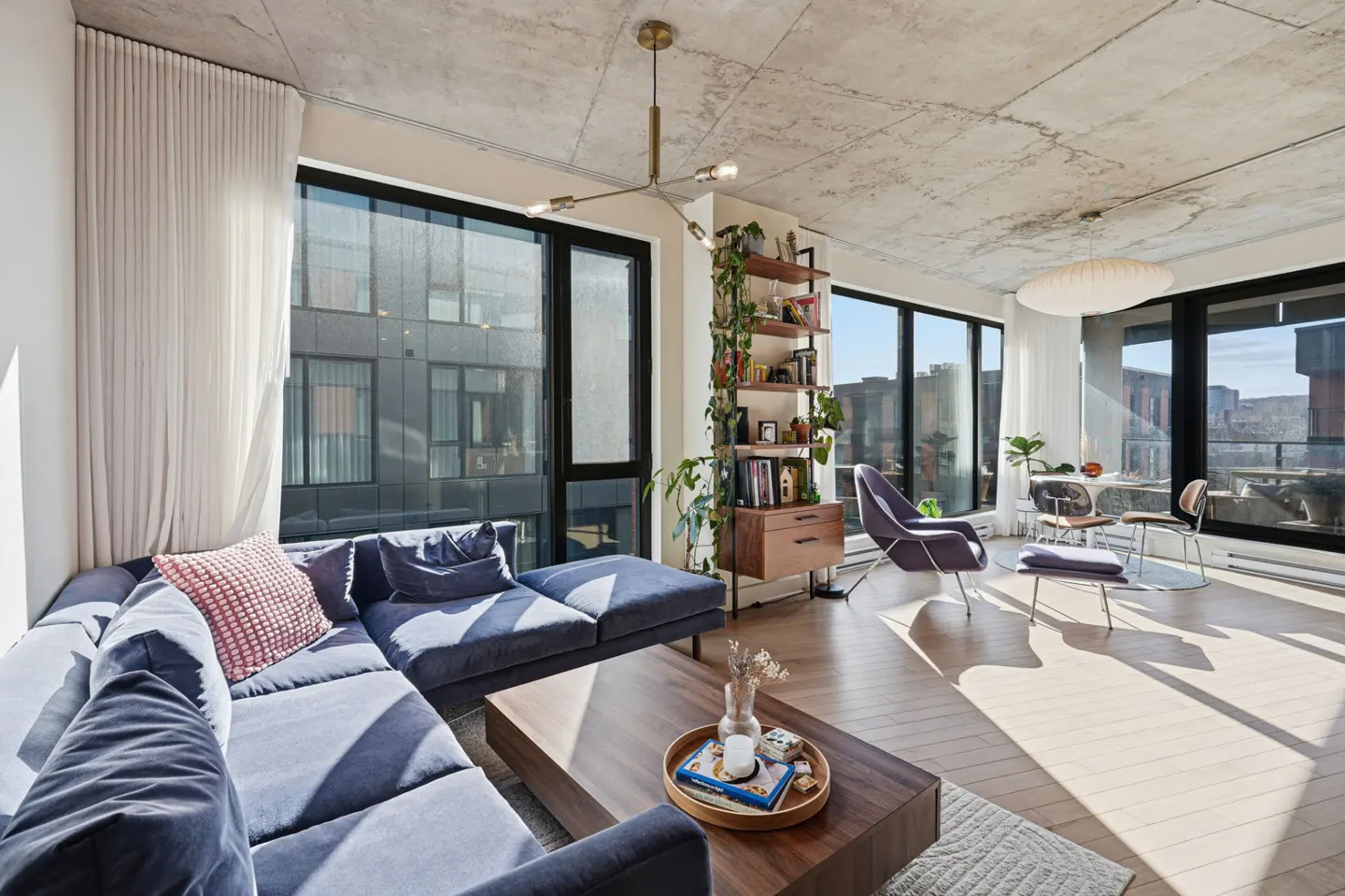 Bright living room with a blue sectional sofa, wood coffee table, and large windows with city views. A bookshelf with plants adds a touch of nature.