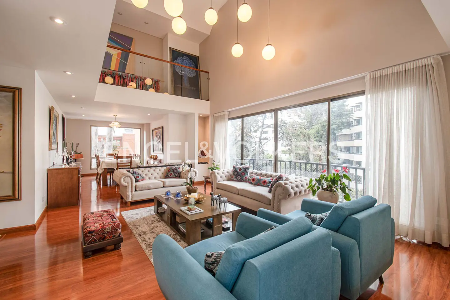 Bright, open living room with hardwood floors, blue chairs, and beige sofas. Large windows offer a view of trees and buildings.