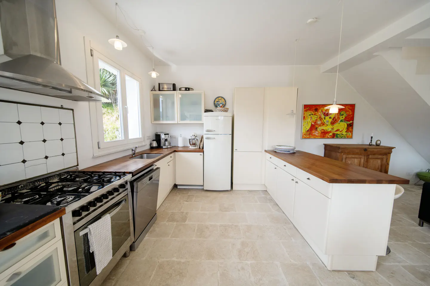 Bright kitchen with white cabinets, wood countertops, and stone tile floor. Stainless steel stove and hood. Colorful painting on the wall.