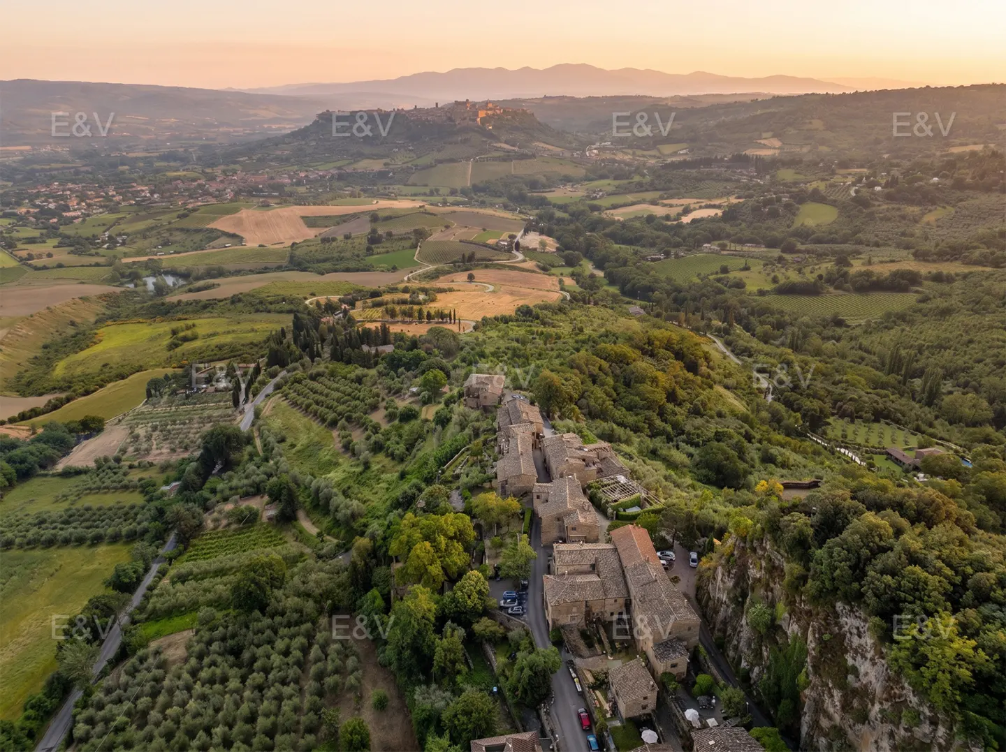 Aerial view of a stone village nestled in a lush, rolling Italian landscape at sunset with distant hills.