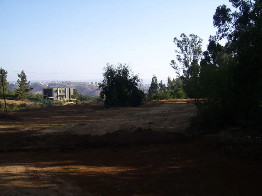 Cleared land for sale, with a modern house in the distance. Trees surround the property, and a city skyline is visible.