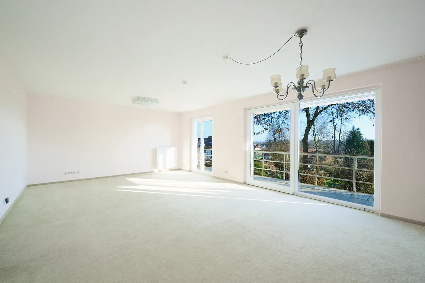 Bright, empty room with light green carpet, white walls, and a chandelier. Large sliding glass doors lead to a balcony with a view of trees.