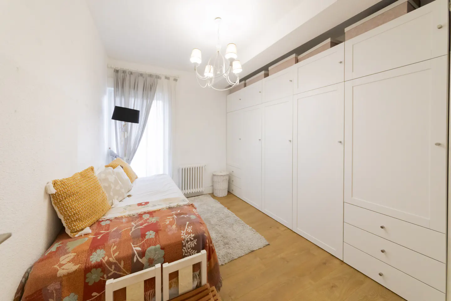 Bedroom with a single bed, orange floral blanket, white walls, and a large white wardrobe. A chandelier hangs from the ceiling.