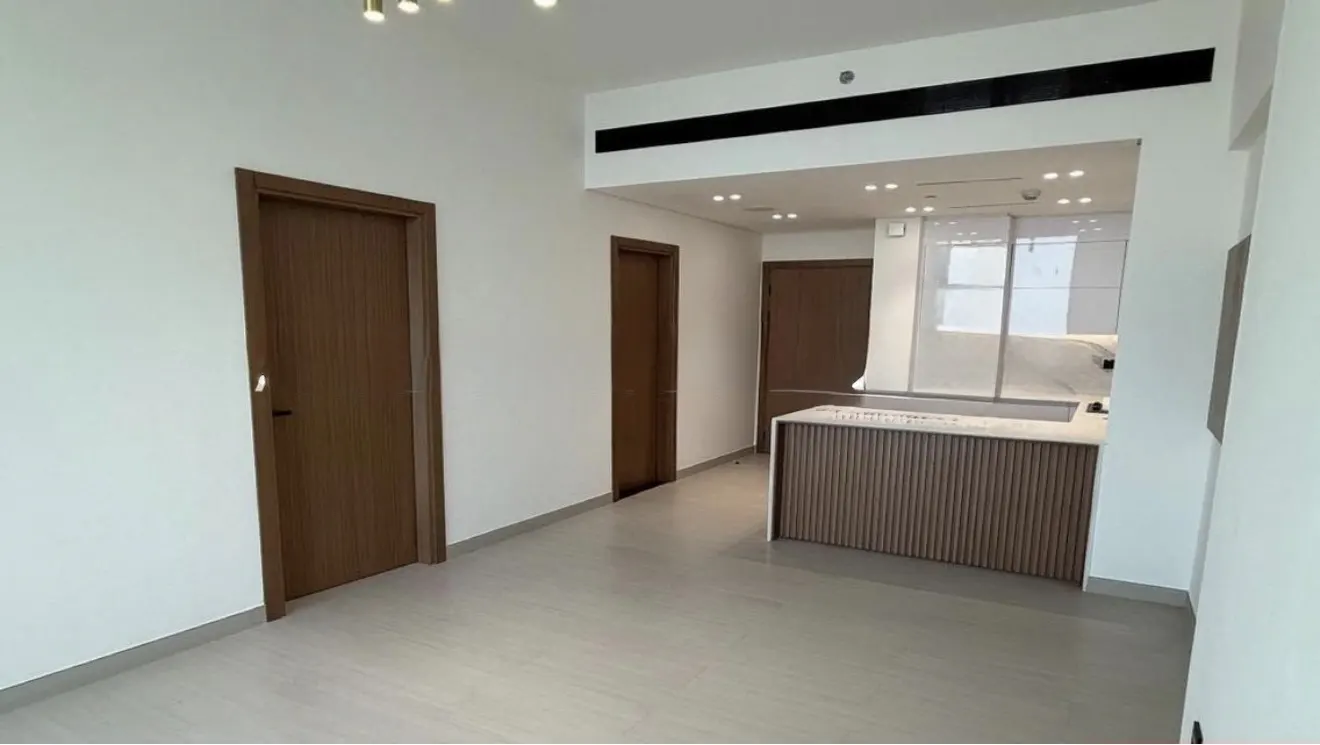An interior shot of a modern apartment with white walls, light gray floors, and brown wooden doors. A kitchen island with a white countertop is visible.
