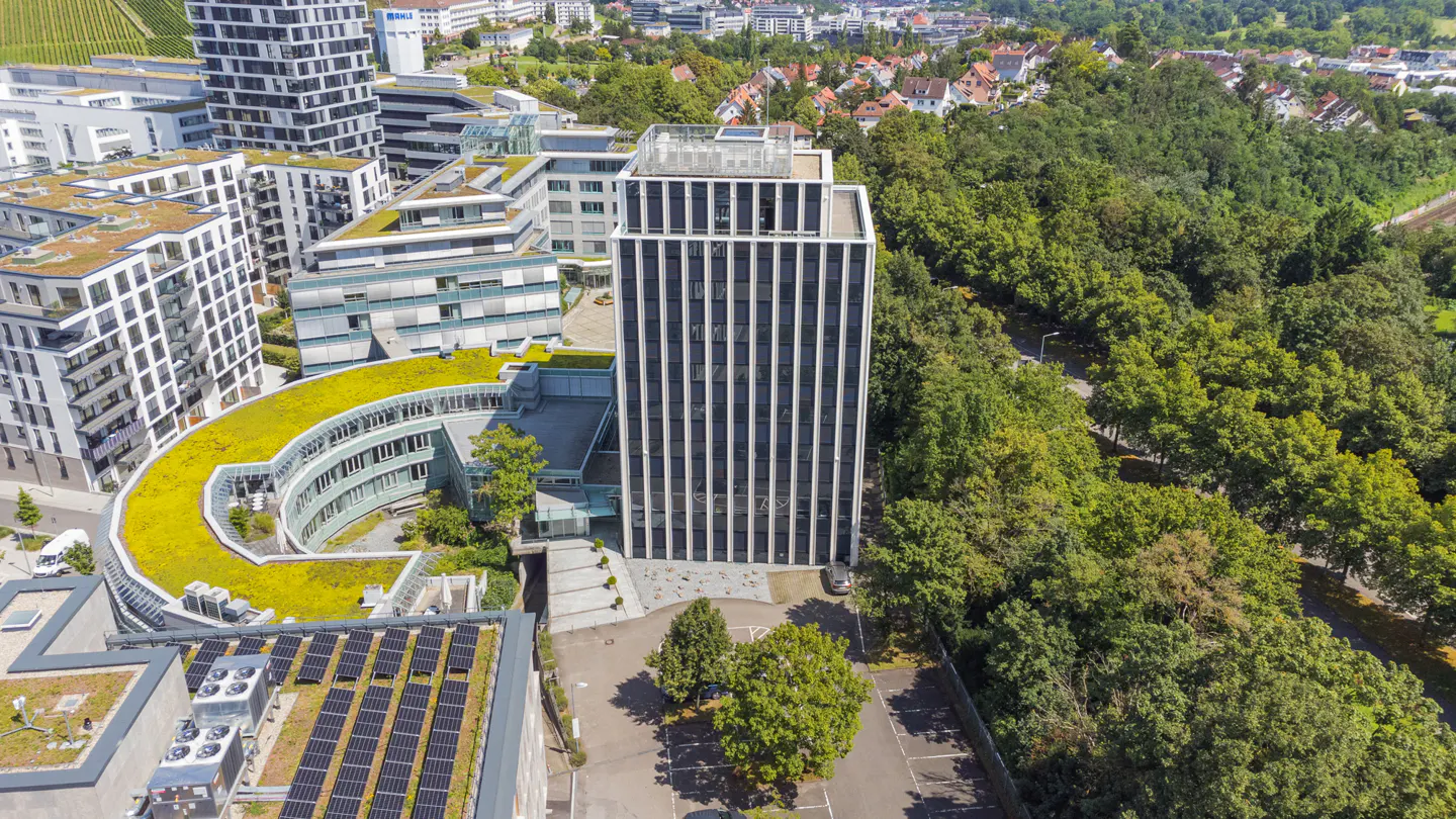 Aerial view of a modern office building with green roofs, solar panels, and a tall, striped skyscraper next to a lush green forest.