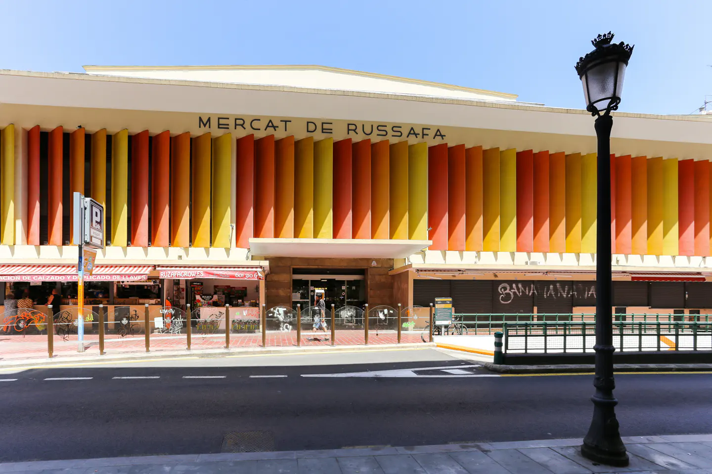 Exterior view of Mercat de Russafa, Valencia, with orange and yellow vertical panels. A black lamppost stands on the right.