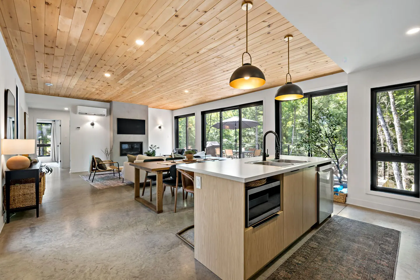 Modern open-concept kitchen and living area with wood-paneled ceiling, concrete floors, and large windows overlooking trees.