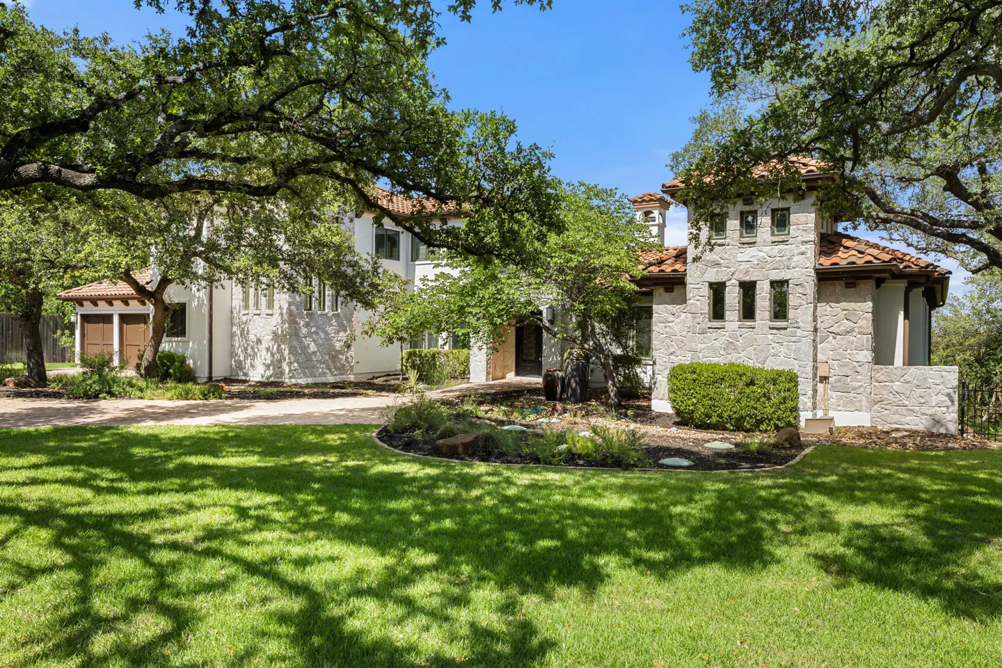 A two-story stone and stucco home with a terracotta roof, surrounded by large oak trees and a lush green lawn.