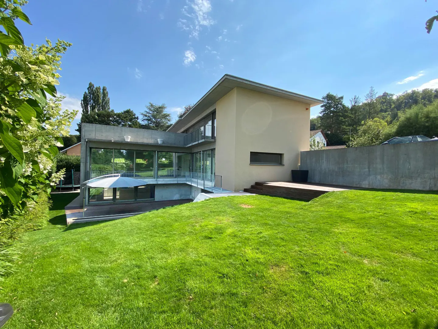 Modern two-story home with large glass windows, a concrete wall, and a spacious green lawn under a clear blue sky.