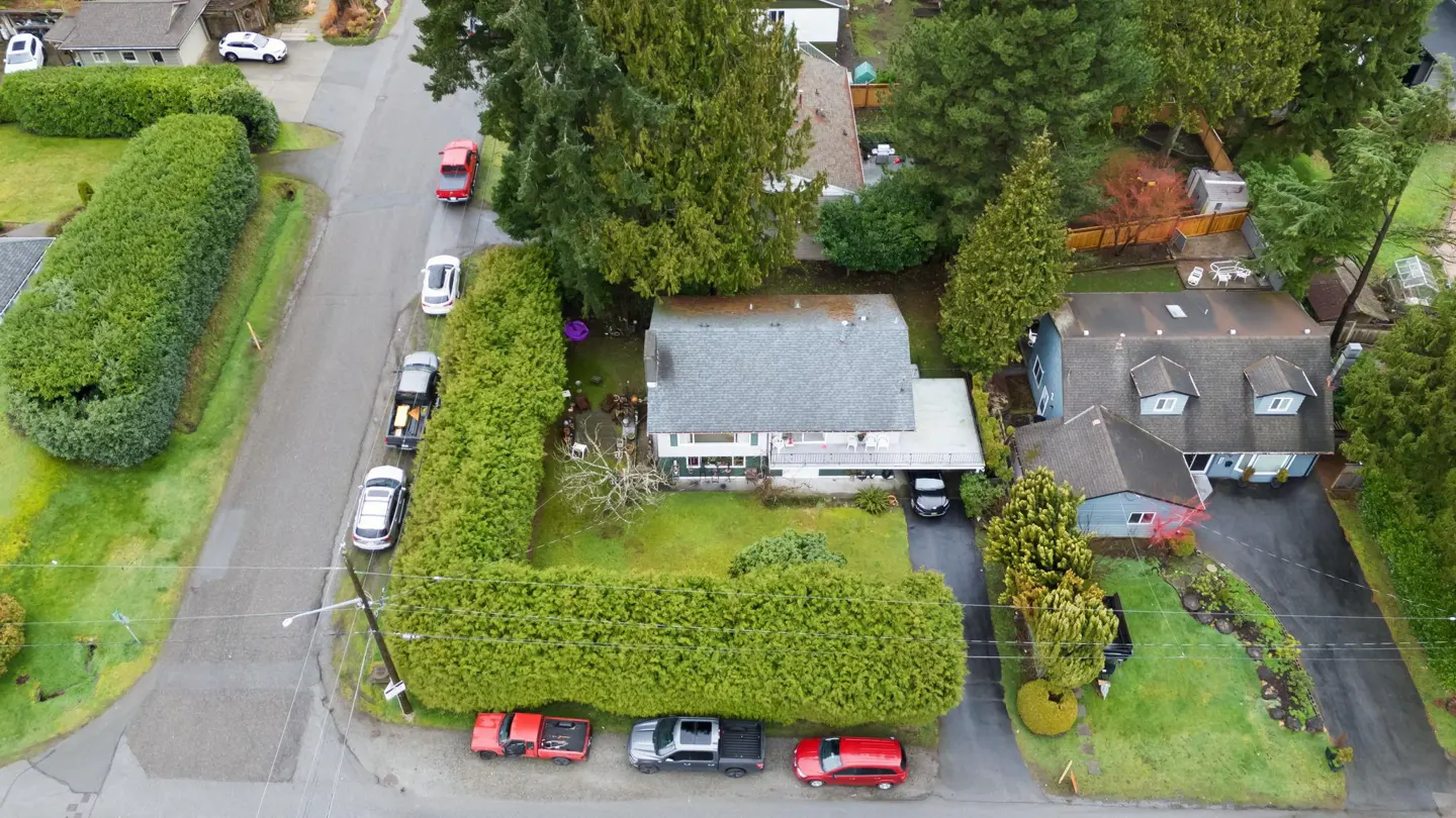 Aerial view of a two-story house with a gray roof, surrounded by tall, green hedges and trees. Cars are parked on the street.