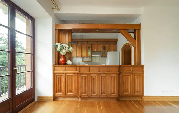 A kitchen with wood cabinets, a bar, and a window. A red vase with white flowers sits on the bar. The floor is wood.