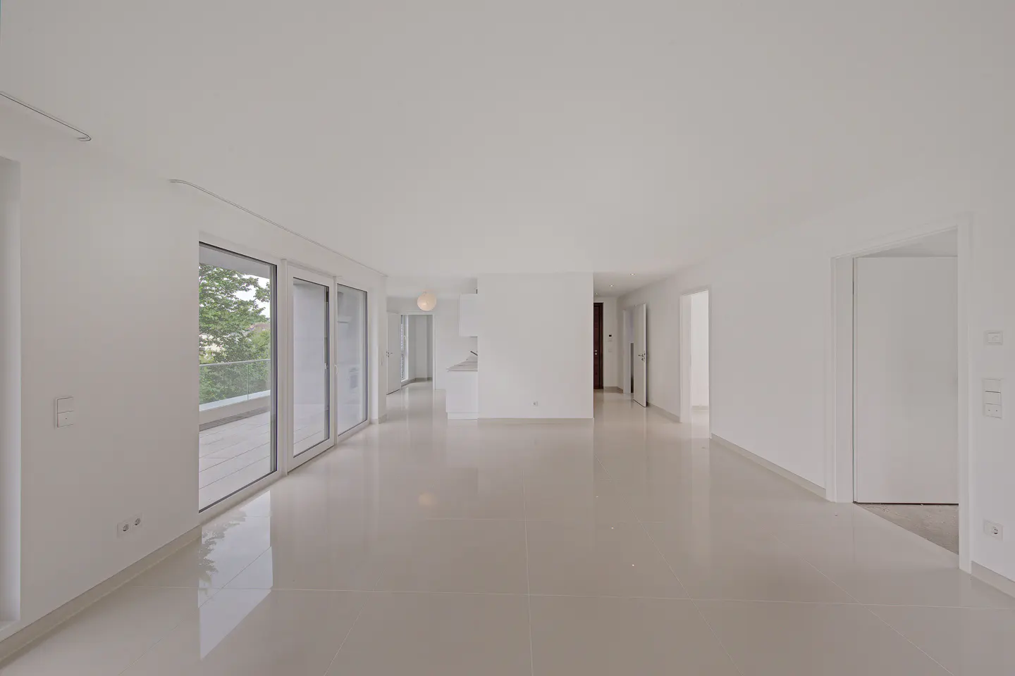 Spacious, minimalist white living room with glossy tile floors, large glass doors to a balcony, and open doorways.