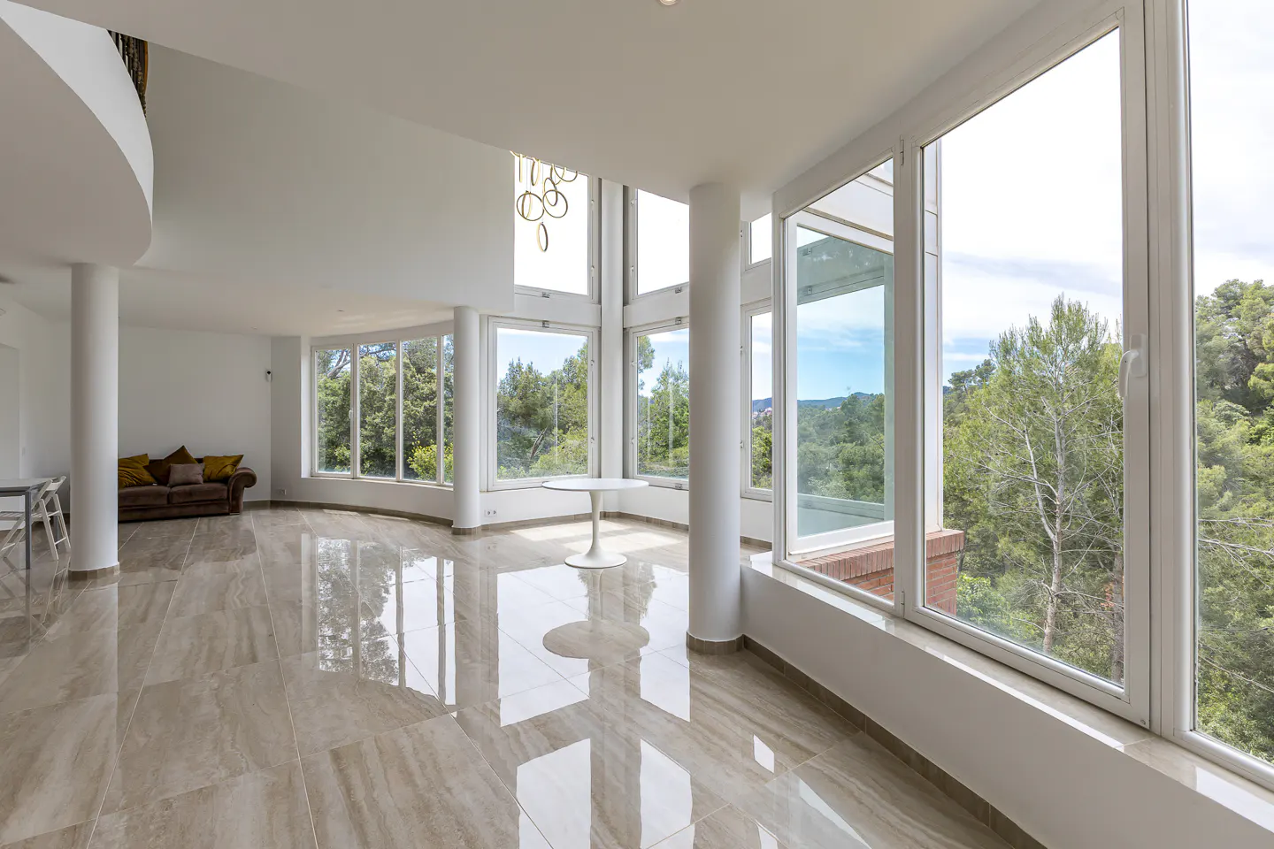 Bright, modern living room with large windows overlooking trees. Beige tile floor reflects light. White walls and columns. Brown sofa in the background.