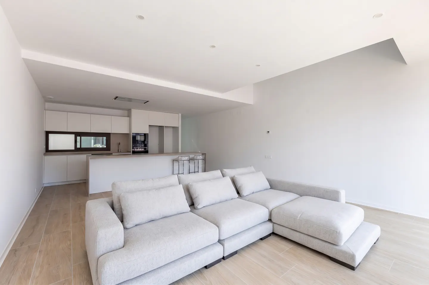 Bright, modern living space with a large, light gray sectional sofa. White kitchen cabinets and island in the background. Wood-look tile flooring.