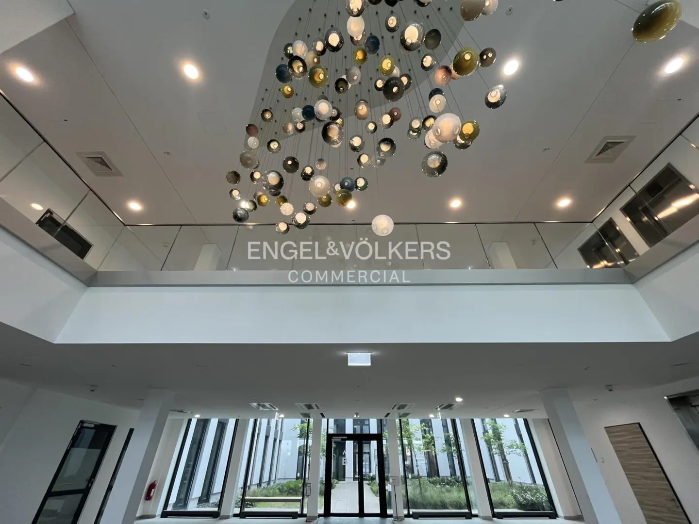 Engel & Völkers Commercial lobby with a modern chandelier. White walls and glass windows offer a view of the outside greenery.