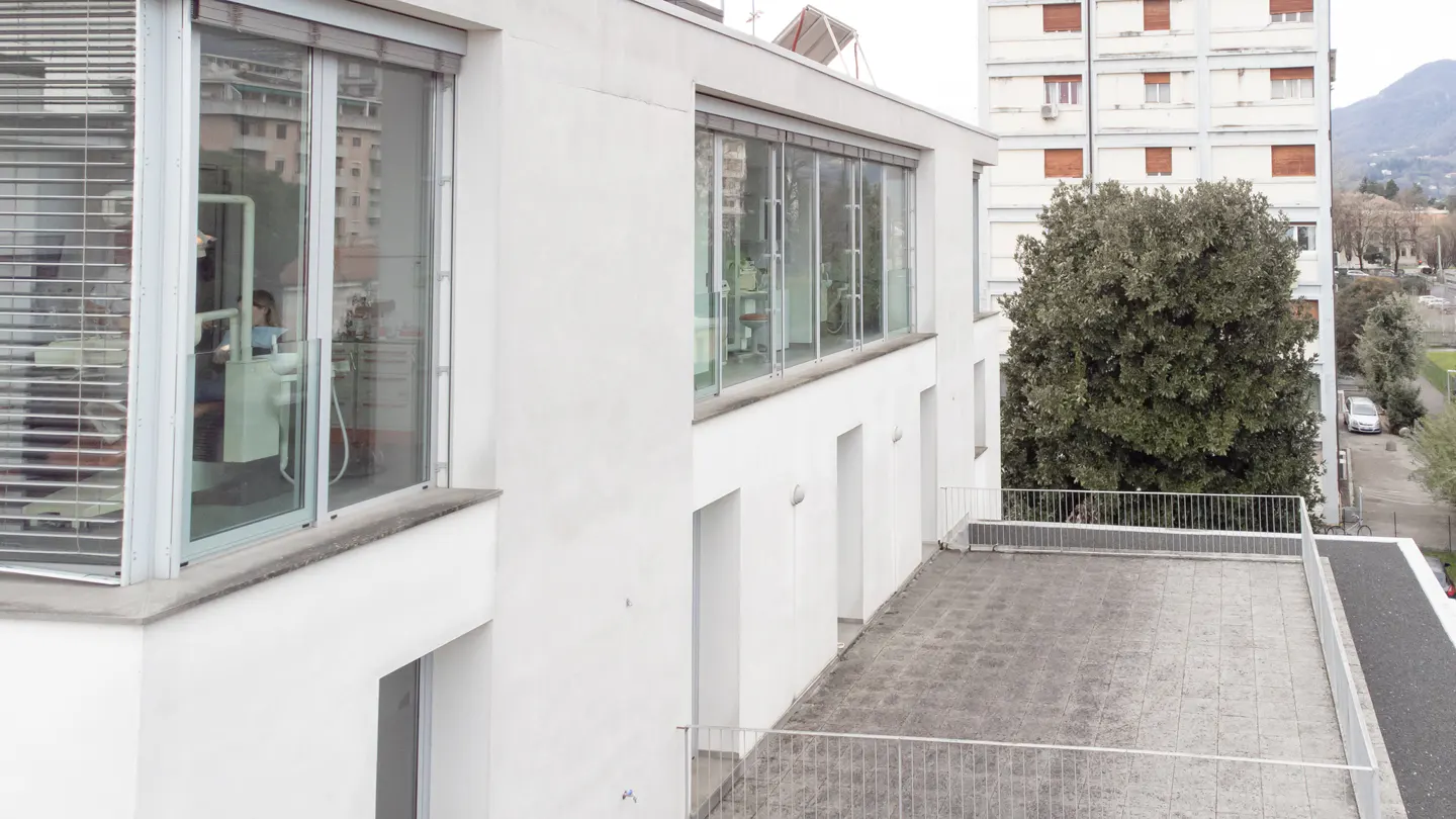 Modern white building exterior with large glass windows, a tiled terrace, and a view of a neighboring apartment building.
