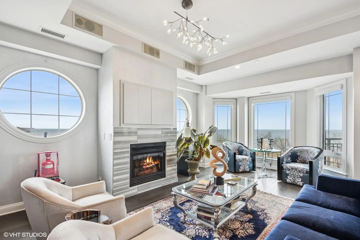Bright living room with a fireplace, round window, and ocean view. Blue sofa and patterned chairs surround a glass coffee table on a blue rug.